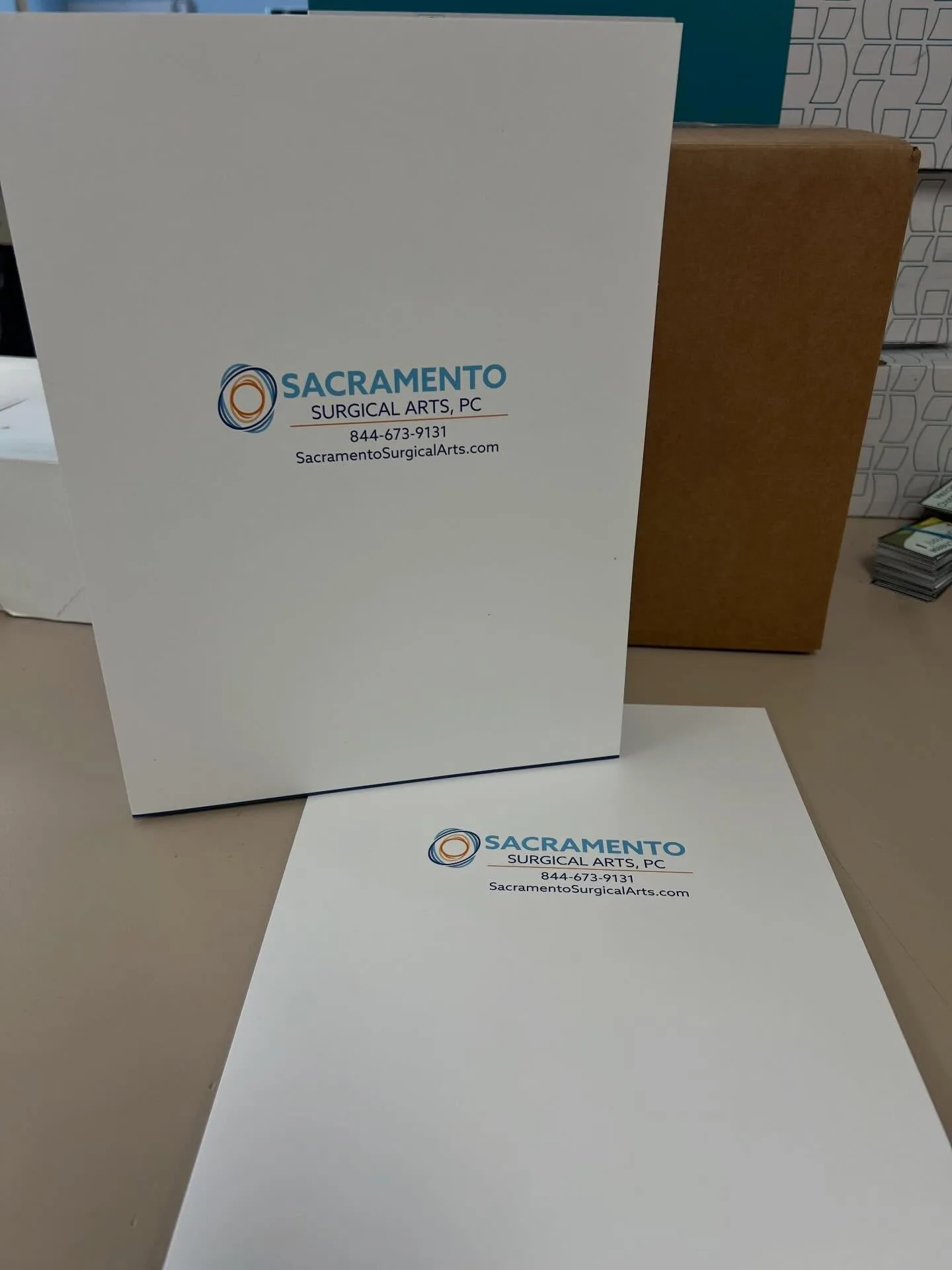 Two printed white presentation folders with the Sacramento Surgical Arts, PC logo, phone number, and website displayed on a counter beside