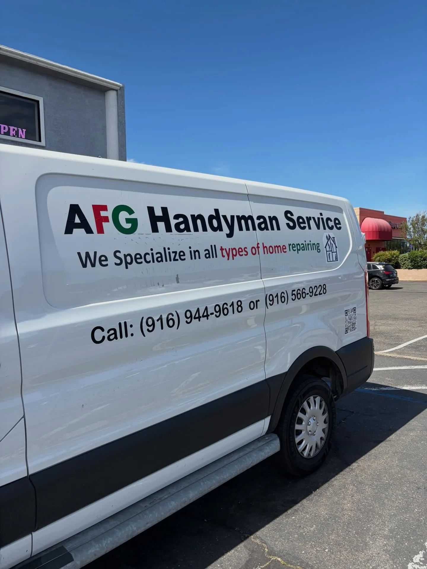 Side view of a white Ford Transit work van with AFG Handyman Service vinyl lettering, tagline, two phone numbers, and a QR code, parked in