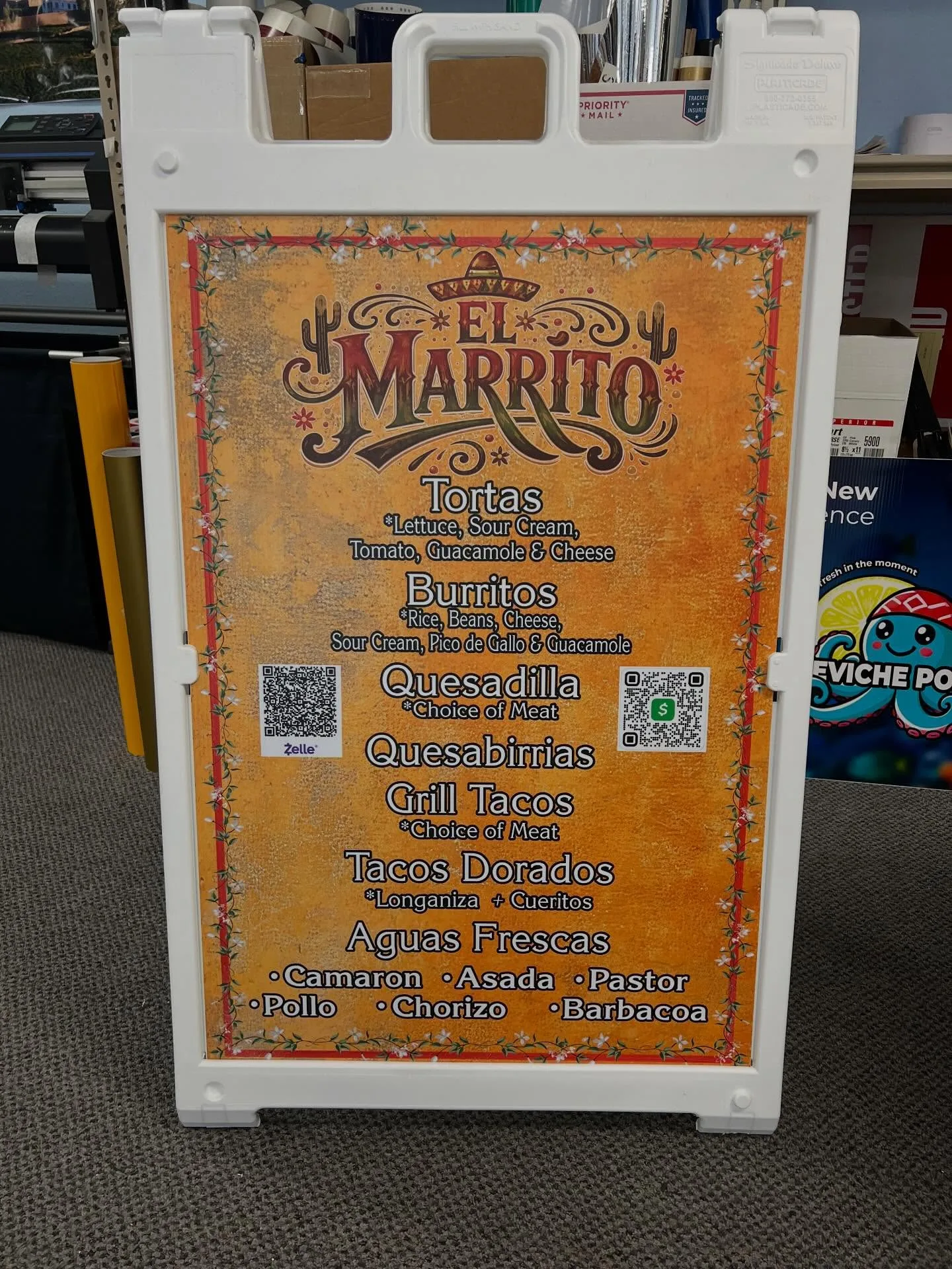 White A-frame sidewalk sign with a full-color printed menu insert for El Marrito taqueria, including QR payment codes, standing on the shop