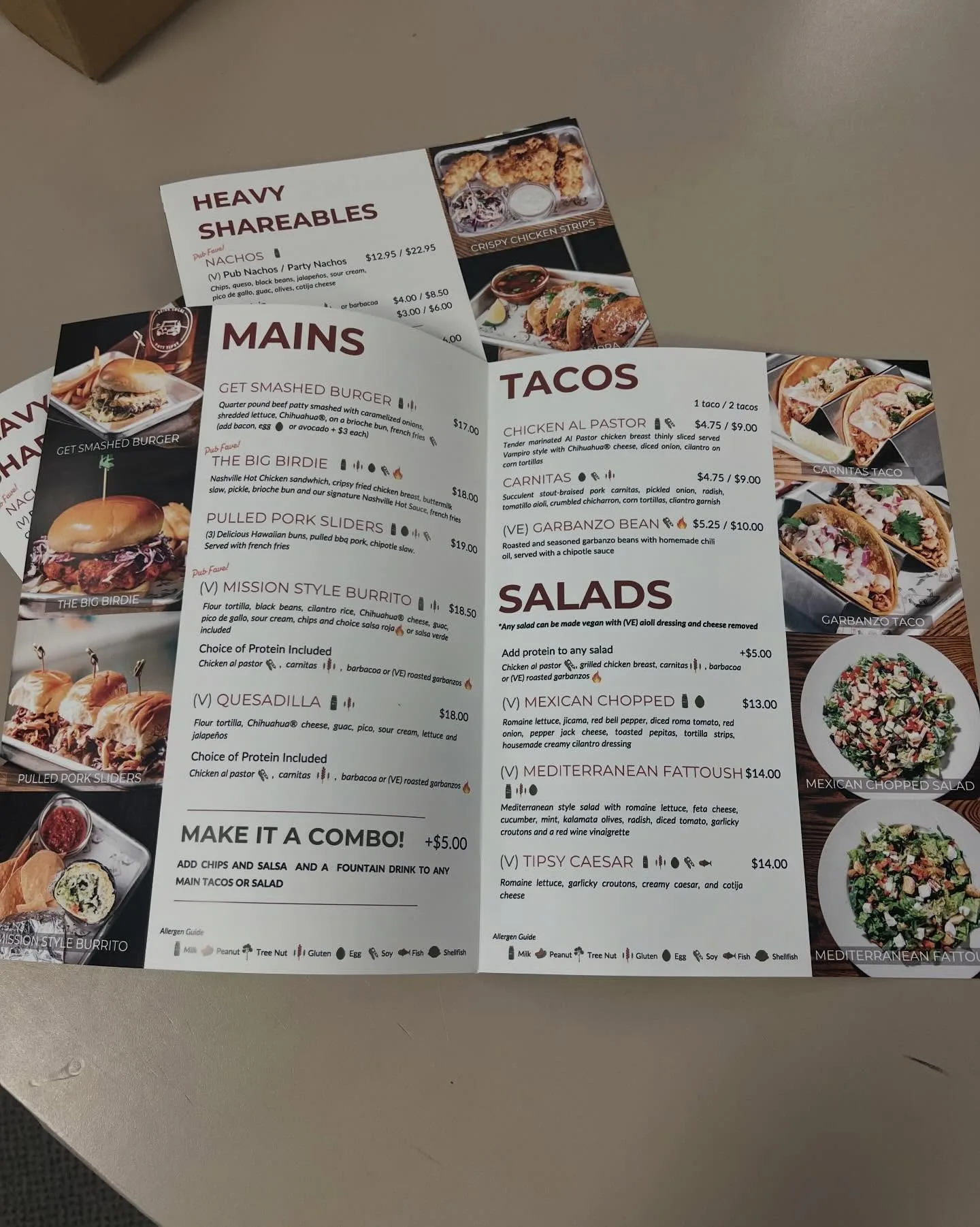 Open printed restaurant menu booklet showing Mains, Tacos, and Salads pages with food photography, lying on a counter with more copies