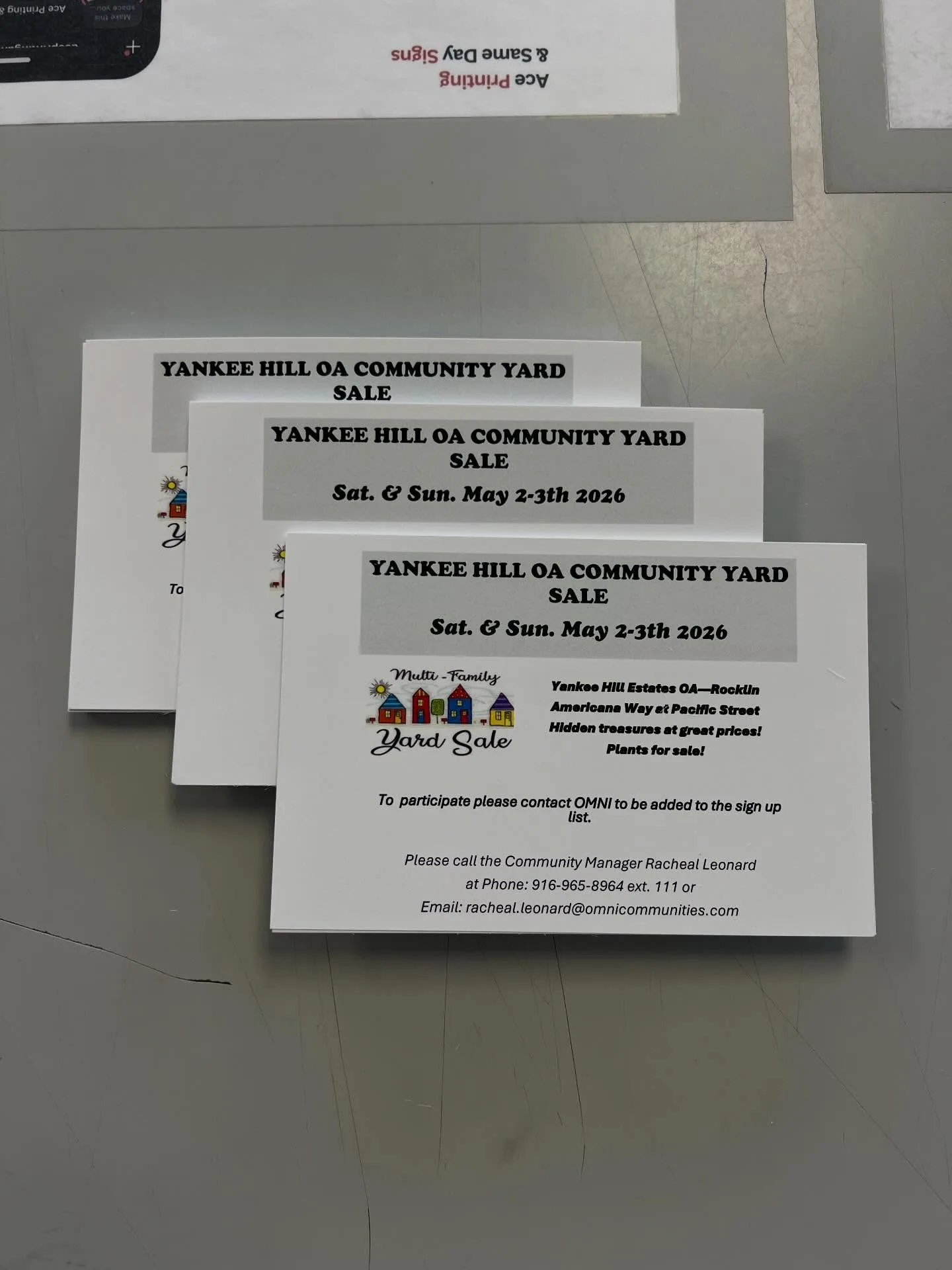 Stacks of printed flyer cards for a 'Yankee Hill OA Community Yard Sale' (Rocklin, May 2-3 2026) laid out on a gray cutting table, with an
