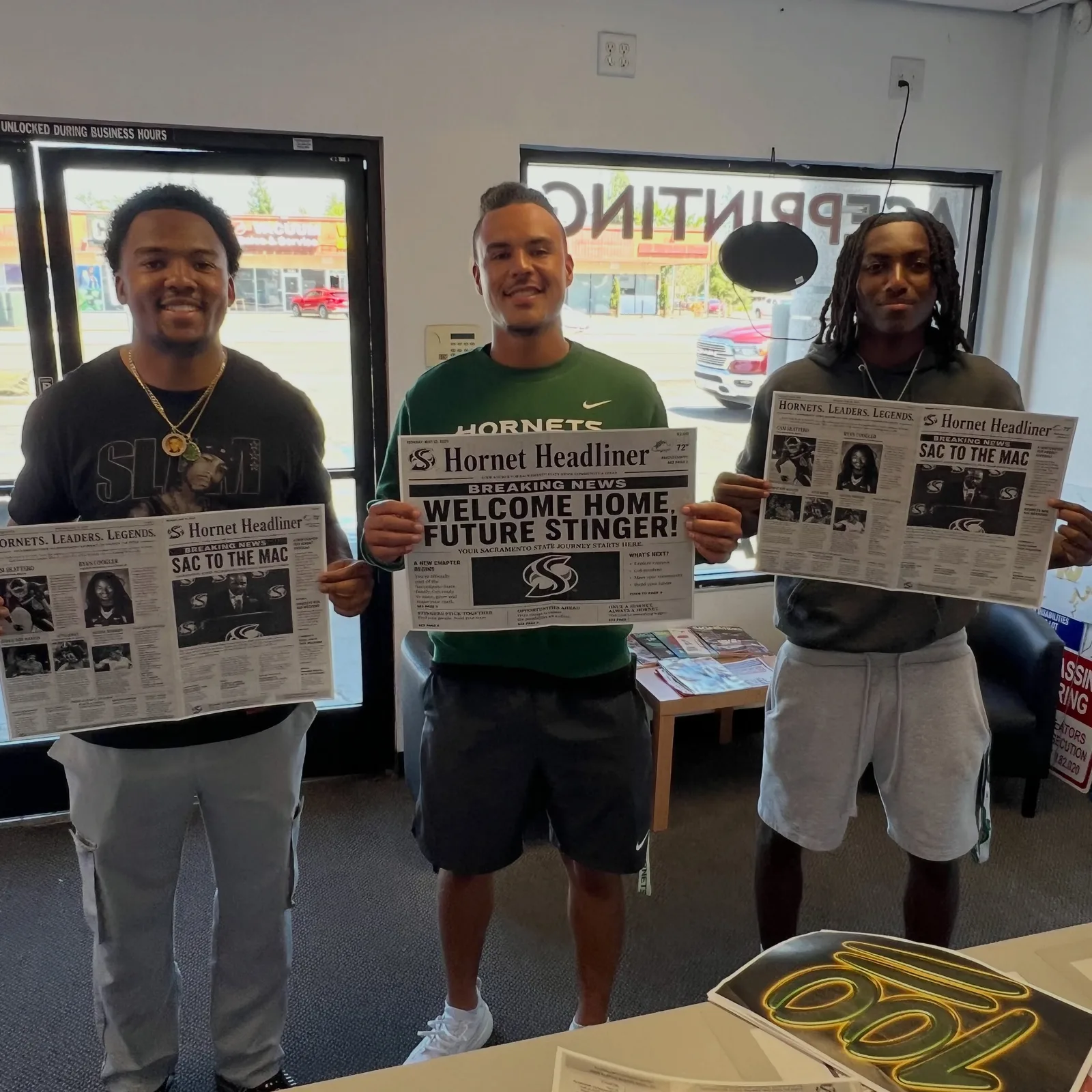 Three smiling young men, one in a Sacramento State Hornets shirt, standing inside the Ace Printing shop holding large custom-printed