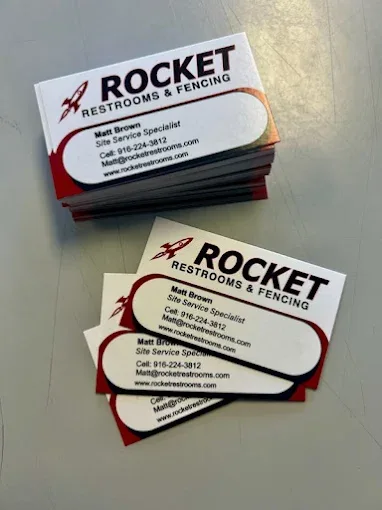 Stacks of freshly printed business cards for Rocket Restrooms & Fencing (Matt Brown, Site Service Specialist) fanned out on a gray table.